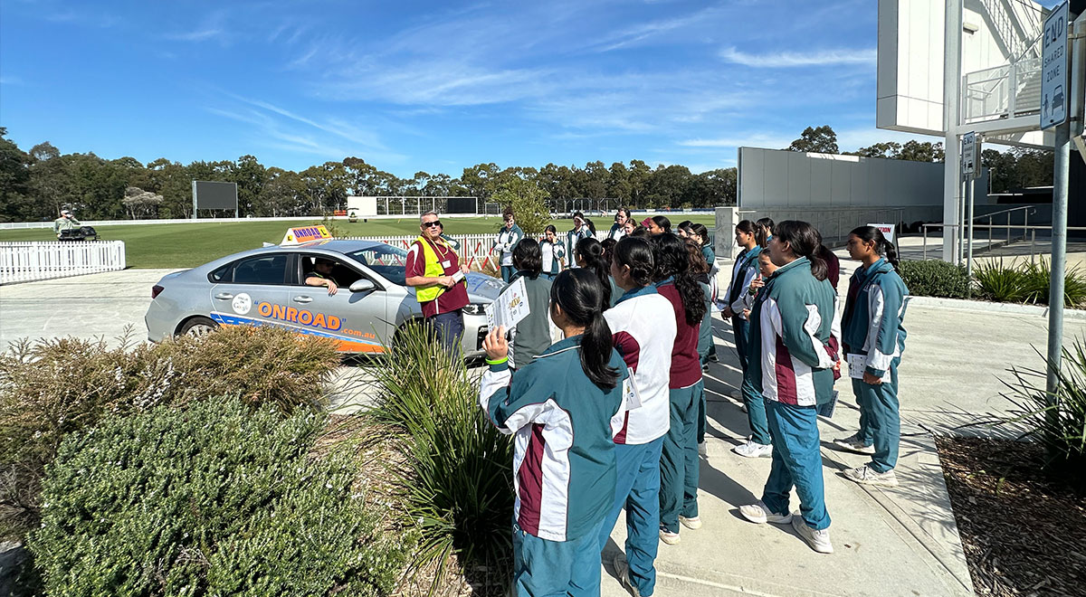 Driving instructor speaking to Catherine McAuley students. The students are crowding around a car
